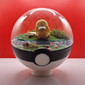 Puede incluir: Una figura amarilla de Psyduck se encuentra en un paisaje en miniatura dentro de un recipiente esférico transparente. La escena incluye un pequeño arroyo, musgo verde y acentos de hongos rosas. El recipiente está sobre una base de Poké Ball en blanco y negro.