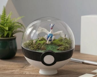 May include: A spherical terrarium shaped like a white and black Poké Ball with a clear top dome. Inside, a white and blue Lugia figurine stands on a patch of green moss and textured ground. The container features a central black band and a white circle.
