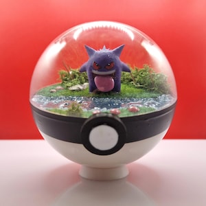 Può includere: Una figura viola di Gengar con una lingua rosa all'interno di un espositore sferico trasparente. La custodia è inserita in una base Pokéball bianca e nera, con un paesaggio in miniatura di erba e acqua. Lo sfondo è rosso.