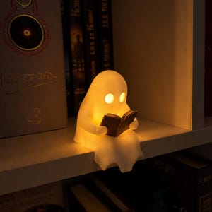 Op de afbeelding: Een kleine, doorschijnende witte lamp in de vorm van een geest die een warm, zacht schijnsel uitstraalt. De geestfiguur zit en houdt een klein, donkerkleurig open boek in de handen. De lamp staat op een plank en verlicht de omgeving.