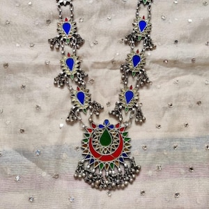 May include: A silver necklace featuring a large, circular pendant with a colourful design. The pendant is made of glass and has red, green, blue and yellow accents. The necklace is decorated with smaller, teardrop-shaped glass pieces in blue and white.