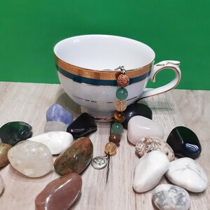 May include: A white teacup with a gold rim and a green and gold band sits on a wooden surface.  A string of colourful beads hangs from the cup's handle.  Surrounding the cup are various smooth, colourful stones.