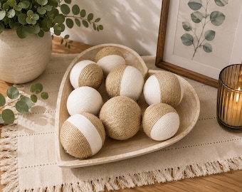 May include: A heart-shaped wooden bowl containing eight decorative spheres. Each sphere is wrapped in a combination of natural tan jute twine and smooth white thread, creating a two-toned, textured appearance within the bowl's light wood finish.