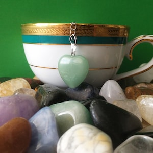 May include: A green heart-shaped tea infuser hangs above a white teacup with gold trim. The cup rests on a bed of colourful polished stones against a green backdrop. The infuser is suspended by a silver chain.