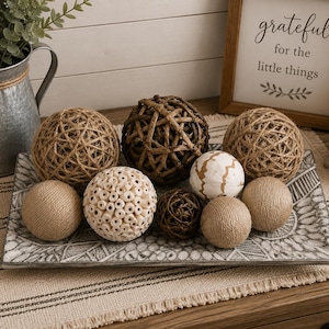 Peut inclure: Un plateau rectangulaire gris avec un motif géométrique blanc contient une collection de sphères décoratives. L'assortiment comprend des boules en jute tressé, une sphère crème texturée avec de petites ouvertures circulaires, une boule en brindilles marron foncé et une boule blanche lisse avec des lignes dorées.