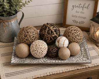 May include: A rectangular gray tray with a white geometric pattern holds a collection of decorative spheres. The assortment includes woven jute balls, a textured cream-colored sphere with small circular openings, a dark brown twig ball, and a smooth white ball with gold lines.