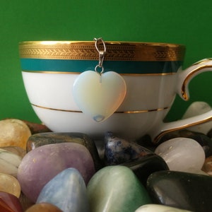 May include: A white porcelain teacup with a gold rim and a green band sits on a bed of colourful stones. A silver chain hangs from the cup, with a white heart-shaped opal pendant.