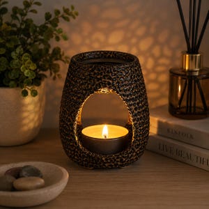 May include: A dark brown ceramic wax warmer with a textured, speckled surface and a central arched opening. A small, round tea light candle sits inside the opening, emitting a warm, glowing light that illuminates the interior of the vessel.