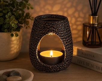 May include: A dark brown ceramic wax warmer with a textured, speckled surface and a central arched opening. A small, round tea light candle sits inside the opening, emitting a warm, glowing light that illuminates the interior of the vessel.