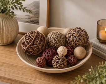 May include: A shallow, light-coloured wooden bowl filled with a collection of decorative spheres. The assortment includes woven rattan balls in brown and dark red, smooth twine-wrapped spheres in beige, and a small, textured cream-coloured ball.
