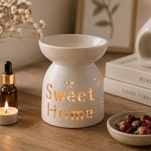 Puede incluir: Un quemador de aceite de cerámica blanca con un recipiente superior ancho y una base redondeada que presenta detalles florales recortados y el texto Sweet Home. El diseño permite que la luz pase a través de las letras y los patrones.