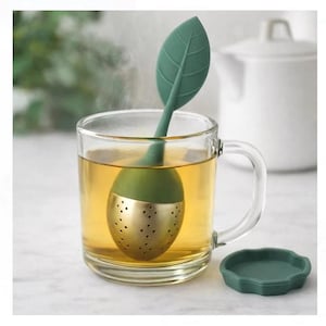 Könnte beinhalten: Ein klares Glas mit Tee, mit einem grünen, blattförmigen Tee-Ei. Das Tee-Ei hat einen goldfarbenen, perforierten unteren Teil. Ein passender grüner Deckel liegt neben der Tasse. Im Hintergrund eine weiße Teekanne.