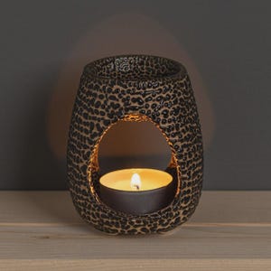 May include: A black and tan ceramic essential oil diffuser with a leopard print design. The diffuser has a large oval opening and a lit tealight candle inside, casting a warm glow. The product is on a wooden surface.