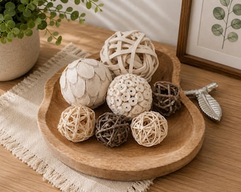 May include: A shallow, organic-shaped wooden bowl containing a collection of decorative spheres. The assortment includes woven rattan balls in light and dark brown, a sphere with a petal-like texture, and a ball with a perforated surface.