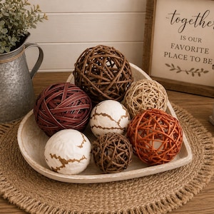 May include: A collection of decorative spheres in a white heart-shaped bowl. The set includes woven rattan balls in dark brown, light tan, and burnt orange, along with smooth white spheres featuring textured, gold-toned crackled patterns.