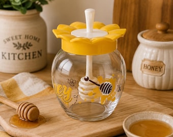 May include: A clear glass honey jar with a yellow sunflower-shaped lid and a white honey dipper. The jar features a graphic print of bees and the word honey in yellow script. The lid has a central white handle for the integrated dipper.