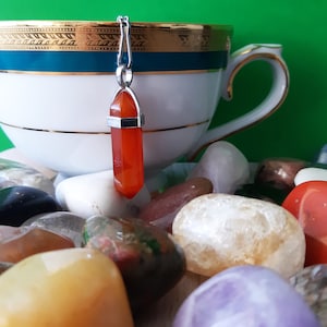 May include: A white teacup with gold trim and a teal band, with a red crystal pendant hanging in front. The cup sits on a bed of colourful polished stones. The background is a solid green.