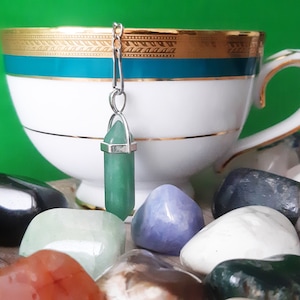 May include: A green gemstone tea infuser suspended in front of a white teacup with gold and teal detailing. The cup is surrounded by a collection of polished stones in various colours, against a green backdrop.