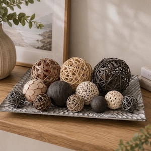 Op de afbeelding: Een rechthoekig grijs dienblad met een gestructureerd geometrisch patroon bevat een verzameling decoratieve bollen in verschillende maten, kleuren en texturen, waaronder gevlochten rotan, glad donkergrijs en lichtgekleurde poreuze ballen.