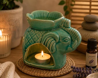May include: A teal ceramic oil burner shaped like an elephant with a lotus flower top. The elephant has a textured back and a hollow centre containing a lit tea light candle. A small brown bottle labelled Lavender Essential Oil sits nearby.