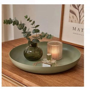 May include: A round, sage green metal tray holding a dark green glass vase with eucalyptus stems, a ribbed glass candle holder with a lit candle, and a small clear glass perfume bottle with a gold cap, arranged on a wooden surface.