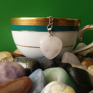 May include: A white teacup with gold trim and a teal band, accompanied by a heart-shaped rose quartz tea infuser. The cup sits on a collection of colourful polished gemstones, against a green background. A stylish tea accessory.