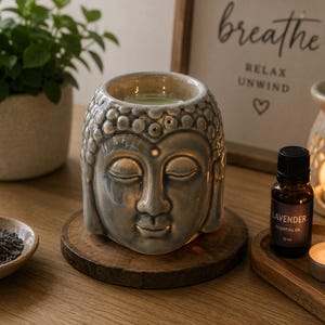 Puede incluir: Un calentador de cera de cerámica gris claro brillante con forma de cabeza de Buda con ojos cerrados y cabello texturizado. La parte superior cuenta con un cuenco empotrado para ceras aromáticas. La pieza descansa sobre una base de madera.