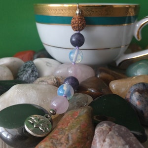 May include: A purple, pink and white crystal beaded charm with a silver charm featuring a lotus flower design. The charm is hanging from a teacup and surrounded by various coloured stones.