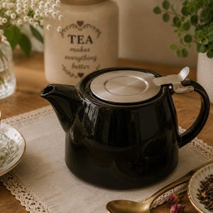 May include: A glossy black ceramic teapot featuring a wide, rounded body, a curved spout, and a sturdy handle. The top is fitted with a circular, brushed stainless steel lid that includes a small, hinged metal tab for easy opening and closing.