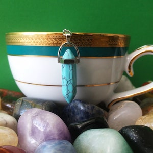 May include: A turquoise crystal pendant with a silver-coloured bail hangs in front of a white teacup with gold trim. The pendant is a light blue colour with dark blue veining. The teacup is surrounded by colourful polished stones.