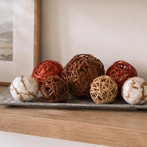 May include: A rectangular gray tray holding a collection of decorative spheres. The arrangement includes two white spheres with gold crackle patterns, three brown woven rattan spheres, and two dark red spheres with a textured, wrapped finish.