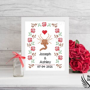 May include: A white framed cross-stitch design with a red heart, a brown alien-like creature, and red roses. The text reads "Joseph & Ashley 07-04-2021".