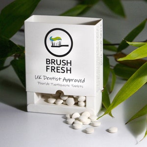 May include: A white box with the text "Brush Fresh" and "UK Dentist Approved Fluoride Toothpaste Tablets". The box is open and contains white toothpaste tablets. Some tablets are scattered on a white surface.