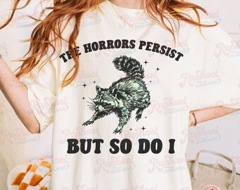 May include: A white t-shirt with a black graphic of a raccoon with the text "The Horrors Persist But So Do I" printed on it.