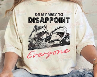 May include: Cream-colored t-shirt featuring a raccoon driving a car, with the text "ON MY WAY TO DISAPPOINT Everyone" in black and red. The graphic is a humorous design, perfect for casual wear.