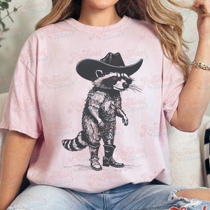 May include: A pink t-shirt with a black and white illustration of a raccoon wearing a cowboy hat and boots. The raccoon is standing on its hind legs and has a serious expression on its face.