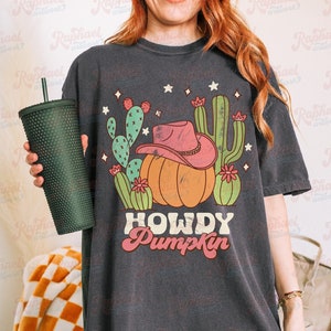 May include: Dark gray t-shirt with a graphic featuring a pumpkin wearing a pink cowboy hat, surrounded by cacti and stars. The text "HOWDY Pumpkin" is below the pumpkin. A dark green tumbler is held in the left hand.
