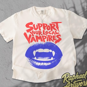 May include: An off-white short-sleeve t-shirt featuring the red text Support Your Local Vampires above a graphic of blue lips with white fangs. The shirt has a crew neckline and a small tag at the collar. The design is centered on the chest.