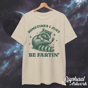 Pode incluir: Uma camiseta bege com um gr&aacute;fico de guaxinim verde e o texto "Sometimes I just be fartin'" impresso na frente.
