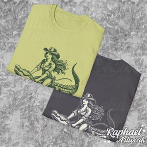 K&ouml;nnte beinhalten: Zwei T-Shirts mit einem grafischen Aufdruck einer Frau, die auf einem Alligator reitet. Die T-Shirts sind in verschiedenen Farben, eines ist hellgr&uuml;n und das andere dunkelgrau.