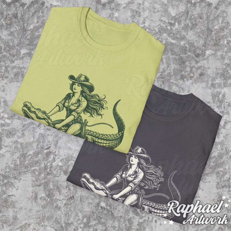 K&ouml;nnte beinhalten: Zwei T-Shirts mit einem grafischen Aufdruck einer Frau, die auf einem Alligator reitet. Die T-Shirts sind in verschiedenen Farben, eines ist hellgr&uuml;n und das andere dunkelgrau.