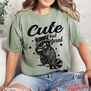 May include: Sage green t-shirt with the text "Cute but feral" above a black and white raccoon graphic eating a hot dog. The design includes black star accents.