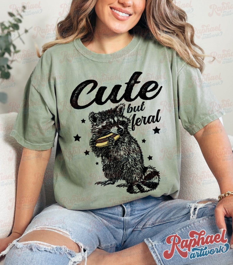 May include: Sage green t-shirt with the text "Cute but feral" above a black and white raccoon graphic eating a hot dog. The design includes black star accents.