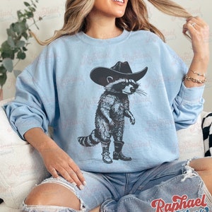 May include: A light blue sweatshirt with a graphic of a raccoon wearing a cowboy hat and boots. The raccoon is standing on its hind legs and has a serious expression on its face.
