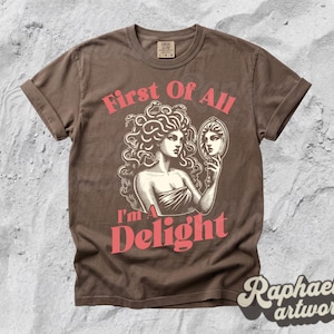 May include: A brown short-sleeve t-shirt featuring a graphic illustration of a figure with snakes for hair holding a hand mirror. The text "First Of All" appears above the figure, and "I'm A Delight" is printed below in a bold, coral-colored font.