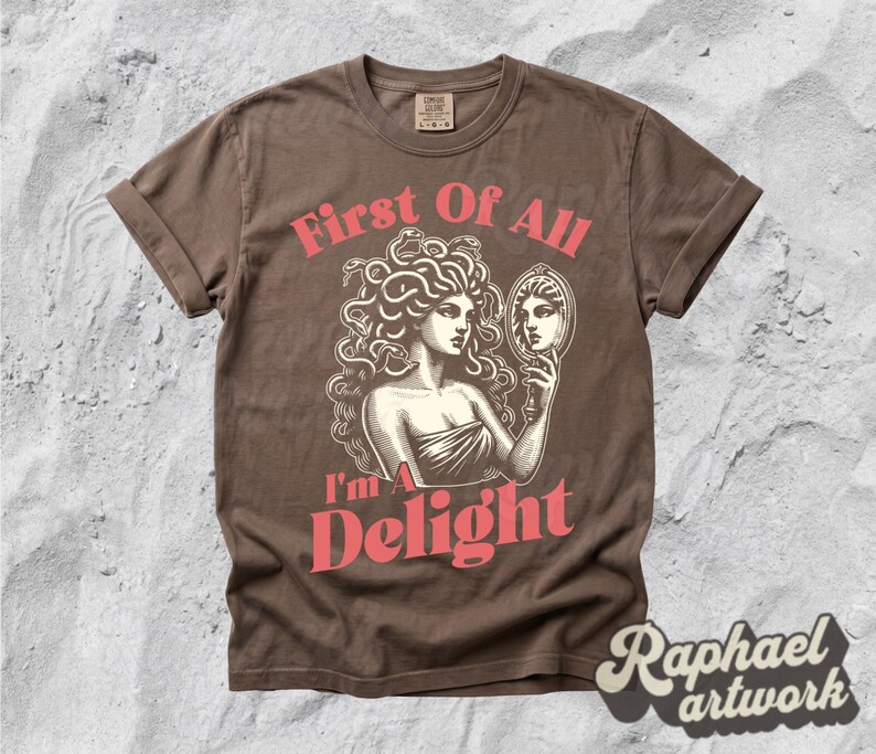 May include: A brown short-sleeve t-shirt featuring a graphic illustration of a figure with snakes for hair holding a hand mirror. The text "First Of All" appears above the figure, and "I'm A Delight" is printed below in a bold, coral-colored font.