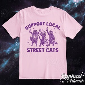 May include: Pink t-shirt with a purple graphic of raccoons and cats drinking beer. The text "Support Local Street Cats" is printed on the shirt.