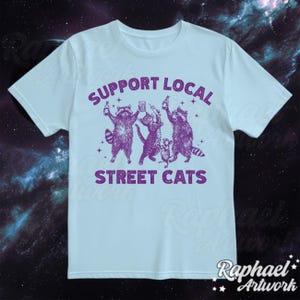 May include: Light blue t-shirt with a purple graphic of raccoons holding drinks and the text "Support Local Street Cats".