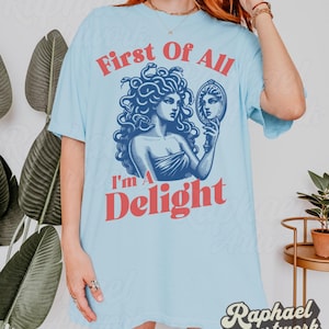 May include: A light blue t-shirt featuring a dark blue graphic of Medusa holding a hand mirror. The text "First Of All" appears in red above the graphic, and the text "I'm A Delight" appears in red below the graphic in a bold, sans-serif font.