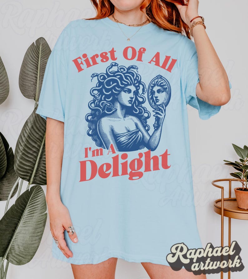May include: A light blue t-shirt featuring a dark blue graphic of Medusa holding a hand mirror. The text "First Of All" appears in red above the graphic, and the text "I'm A Delight" appears in red below the graphic in a bold, sans-serif font.